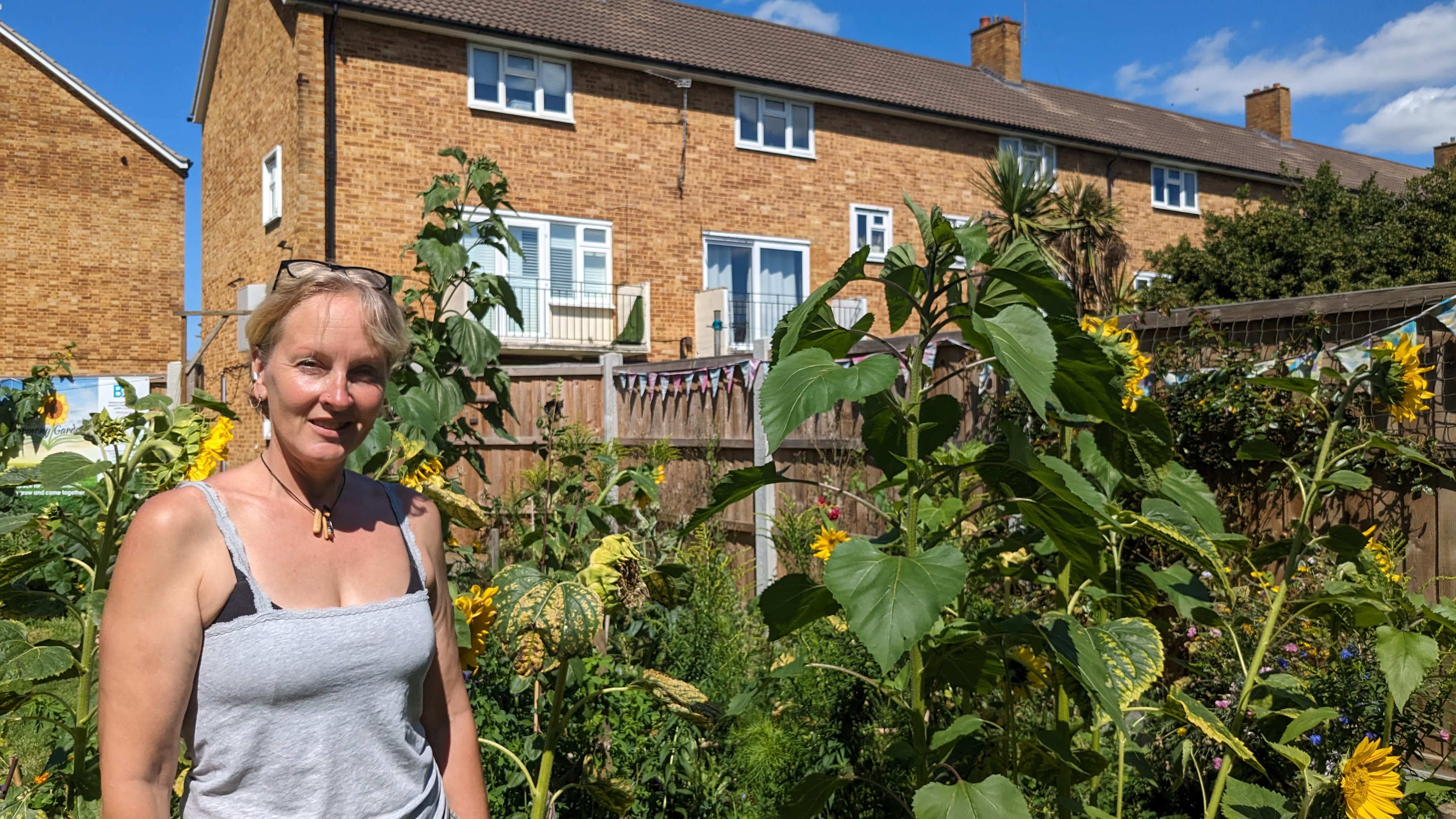 The director of Wyld Edges, a blonde, white woman standing in the garden next to some tall sunflowers. The sun is shining on her, and behind here, there are a few houses
